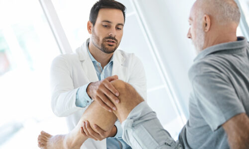 Side view of a doctor in their 30s examining the knee of a senior male patient during a pain management consultation, illustrating real-world context for YPrime’s advanced eCOA solutions that accelerate clinical trials and improve data collection.