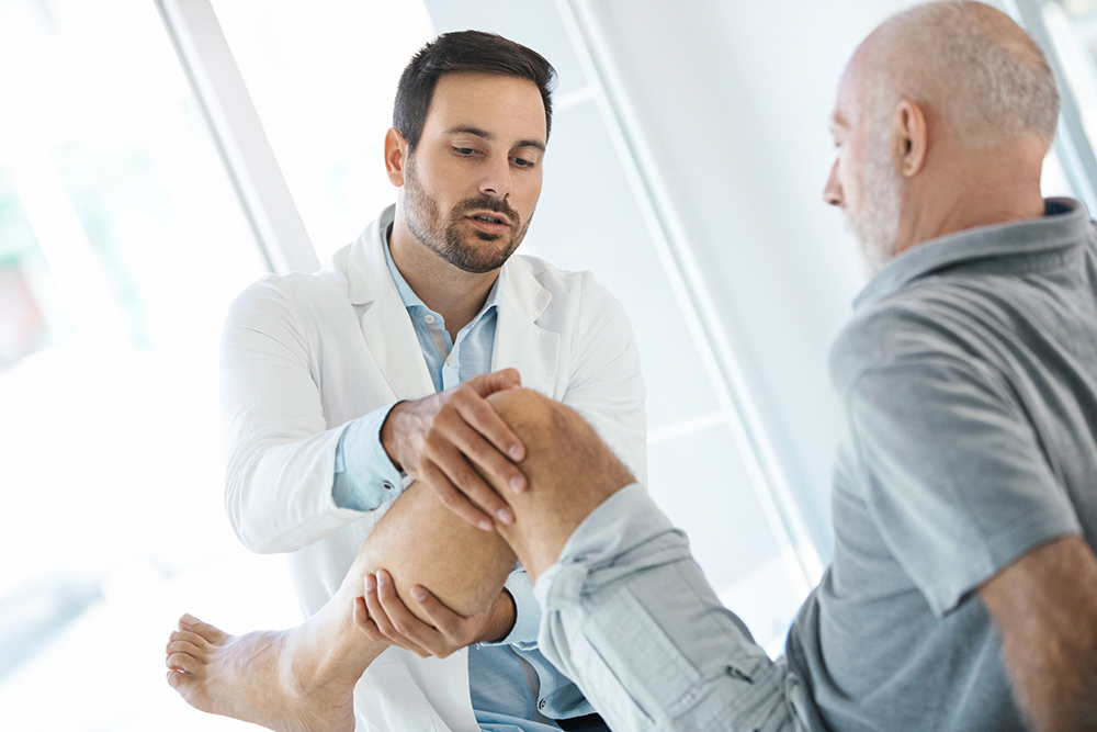 Side view of a doctor in their 30s examining the knee of a senior male patient during a pain management consultation, illustrating real-world context for YPrime’s advanced eCOA solutions that accelerate clinical trials and improve data collection.