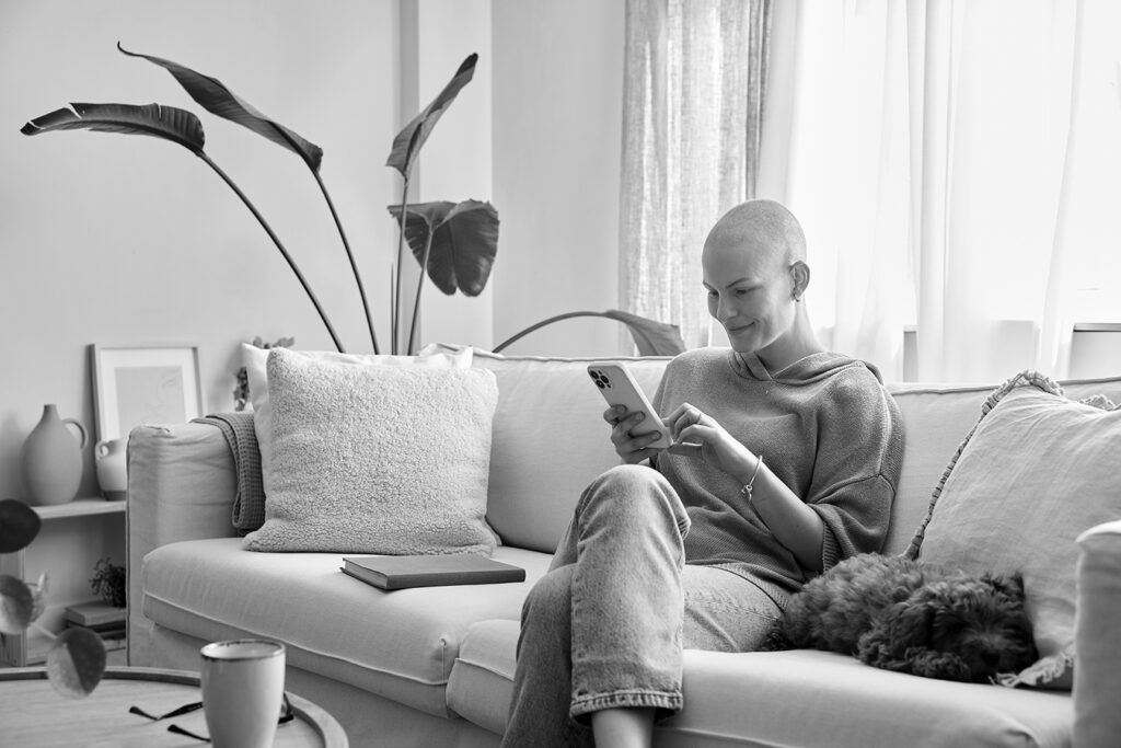 Smiling woman who has lost her hair from cancer treatment sits on a couch with her dog, using her cell phone to access an eCOA platform for oncology clinical trials.
