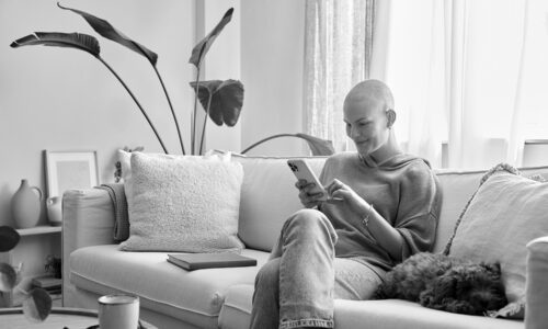 Smiling woman who has lost her hair from cancer treatment sits on a couch with her dog, using her cell phone to access an eCOA platform for oncology clinical trials.